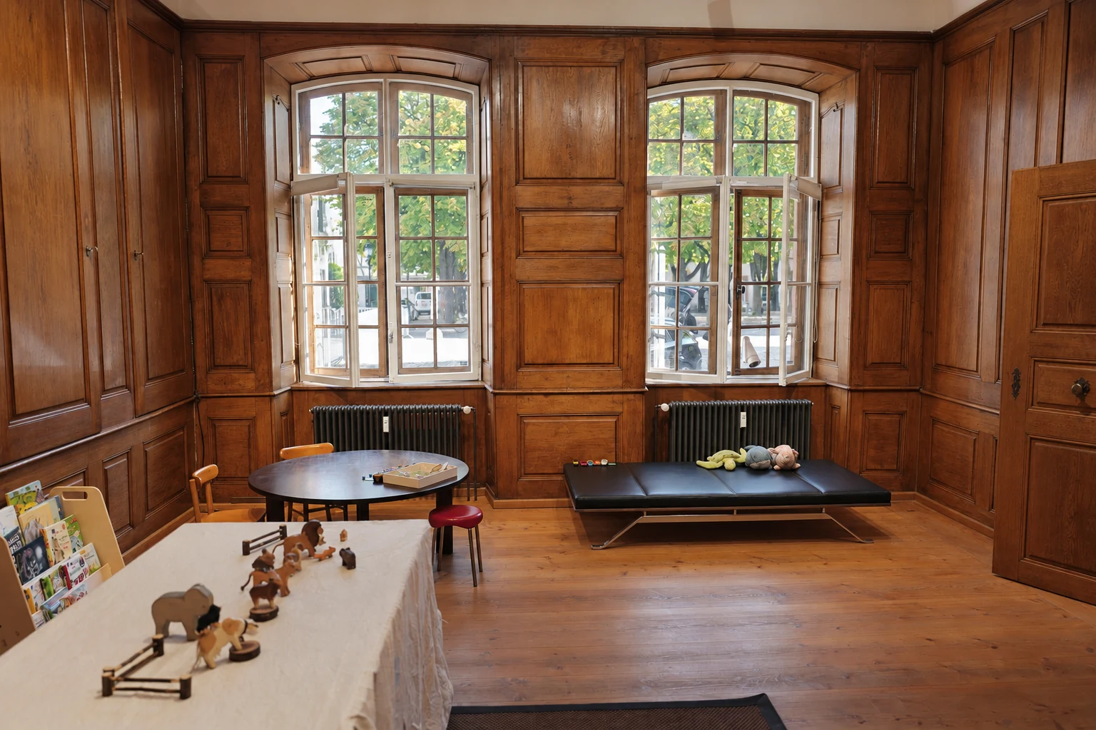 Two large windows in the Lernbus room, with a play table and a padded bench with soft toys.